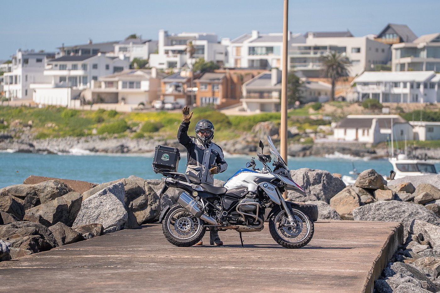 A motorcyclist wearing a helmet, jacket, and gloves waving while standing next to a BMW adventure motorcycle on a dock near water, with rocks, boats, and residential houses in the background. South Africa, Western Cape 