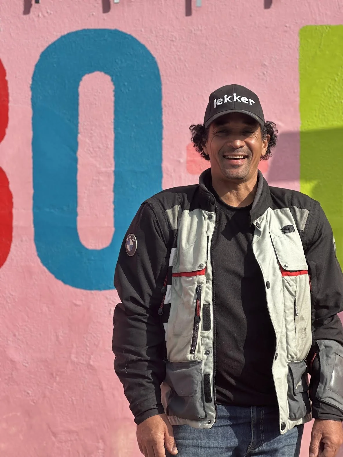 A man standing in front of a colorful mural wall. He is smiling and wearing a black cap with the word "lekker" on it, a black shirt, and a beige and black motorcycle jacket with zipped pockets. Bo Kaap Cape Town South Africa Altitude Adventures Cape