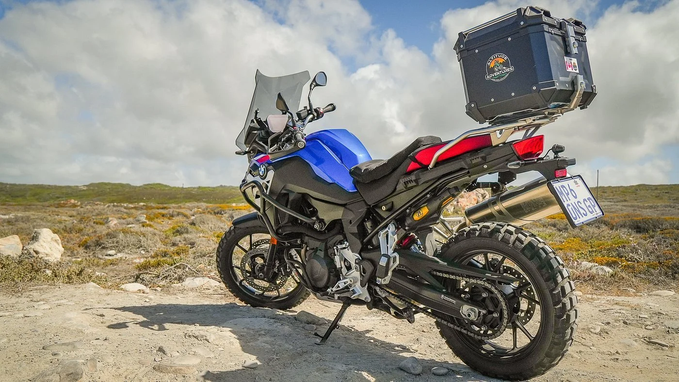 A blue BMW adventure motorcycle with off-road tires parked on a dirt trail in a rugged landscape, carrying a large black storage box on the rear and a fuel tank bag.
