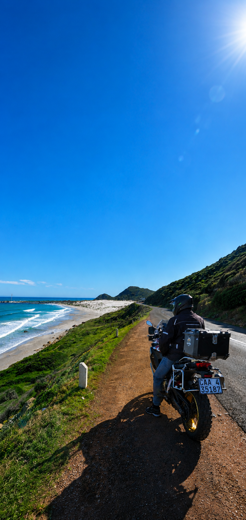 Motorcycle rider wearing a helmet and black jacket with a box on the back, parked on a dirt path next to the ocean, with a grassy hill on the right and a clear blue sky overhead.