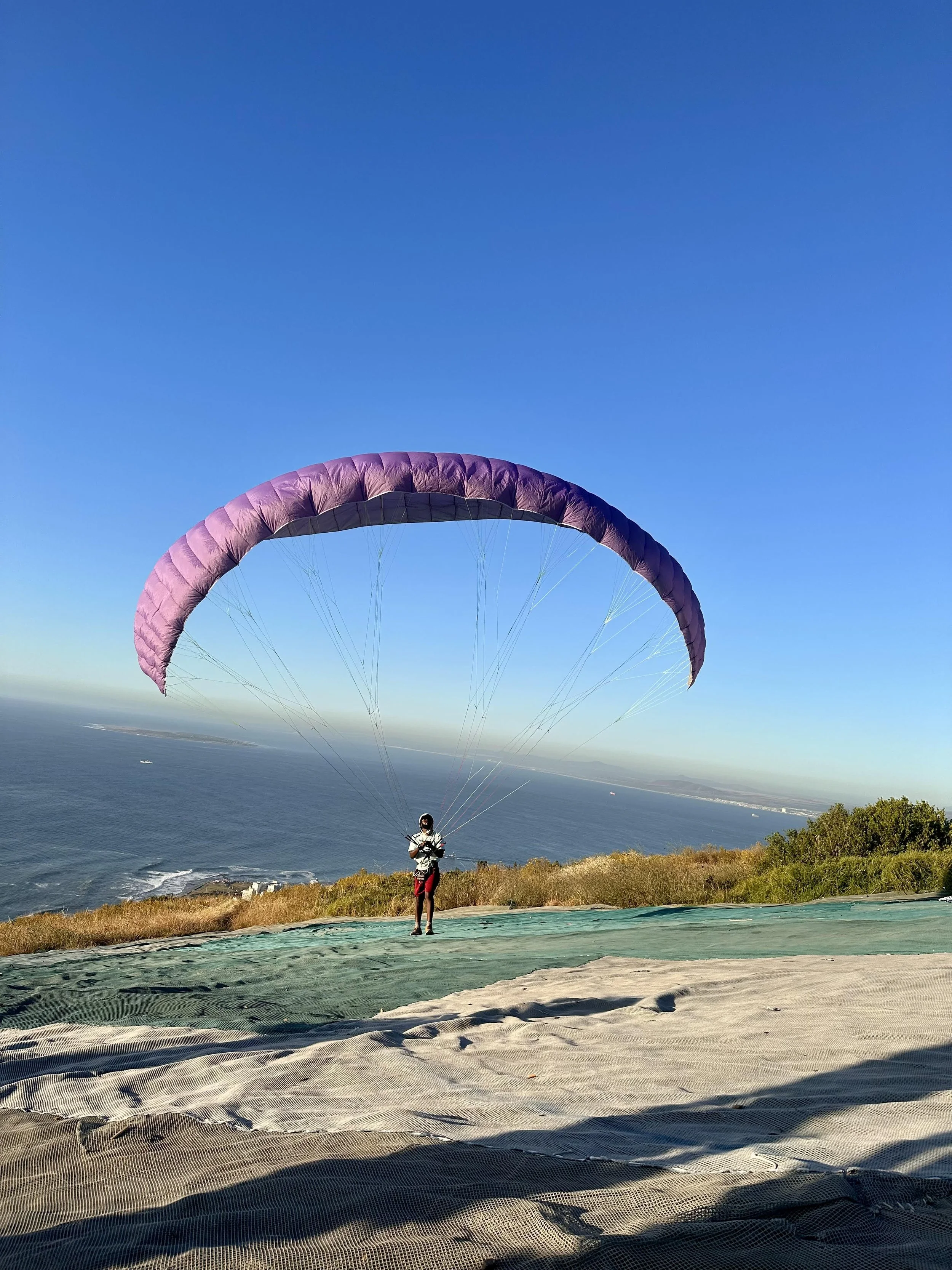 Person preparing to paraglide on a grassy hillside overlooking the ocean under a clear blue sky.