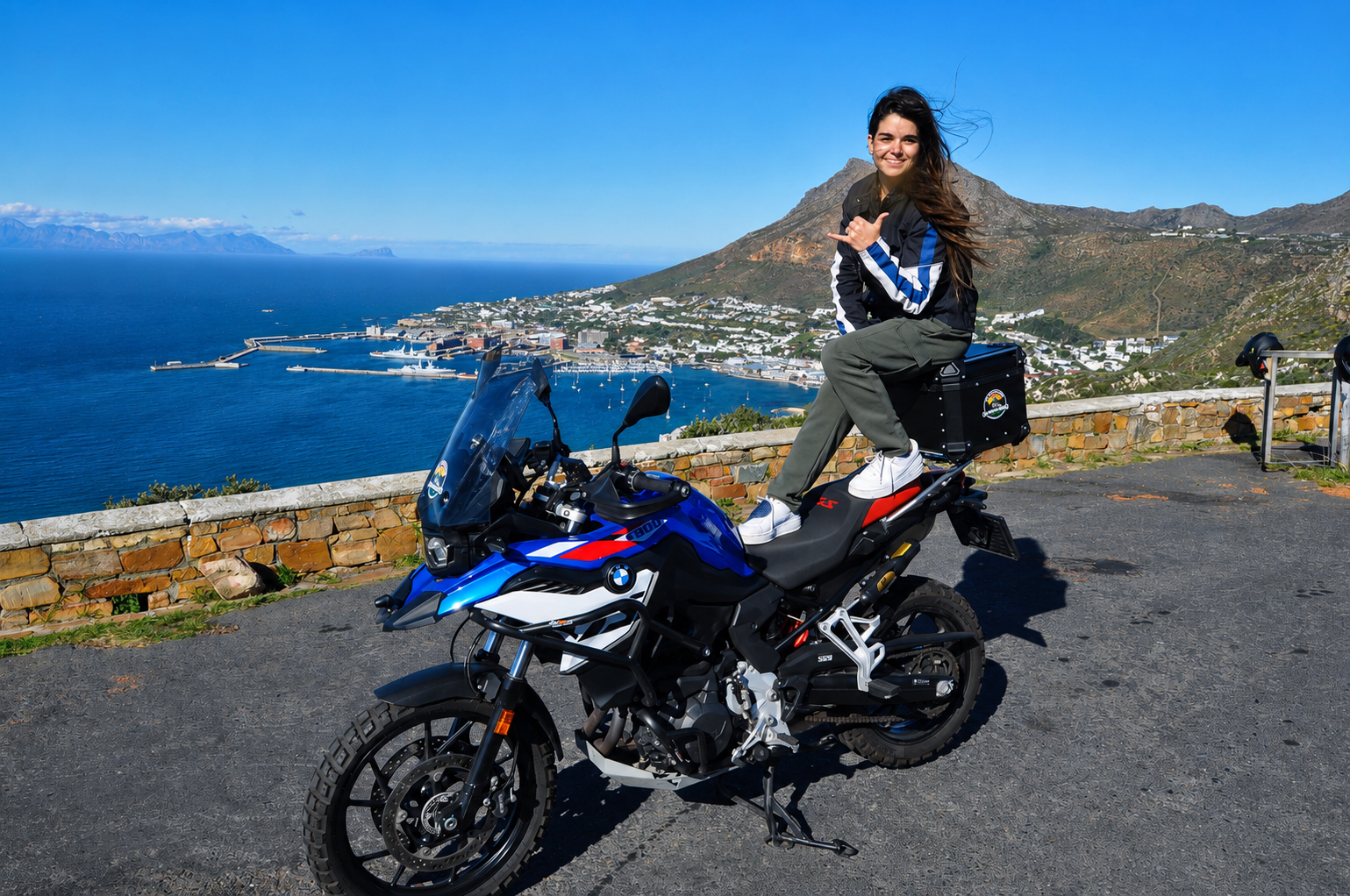 A woman sitting on a blue and black BMW F800 motorcycle with a scenic coastal view of a harbor, mountains, and a clear blue sky in the background. Red Hill Simon's Town Cape Town Southern Suburbs WC South Africa.