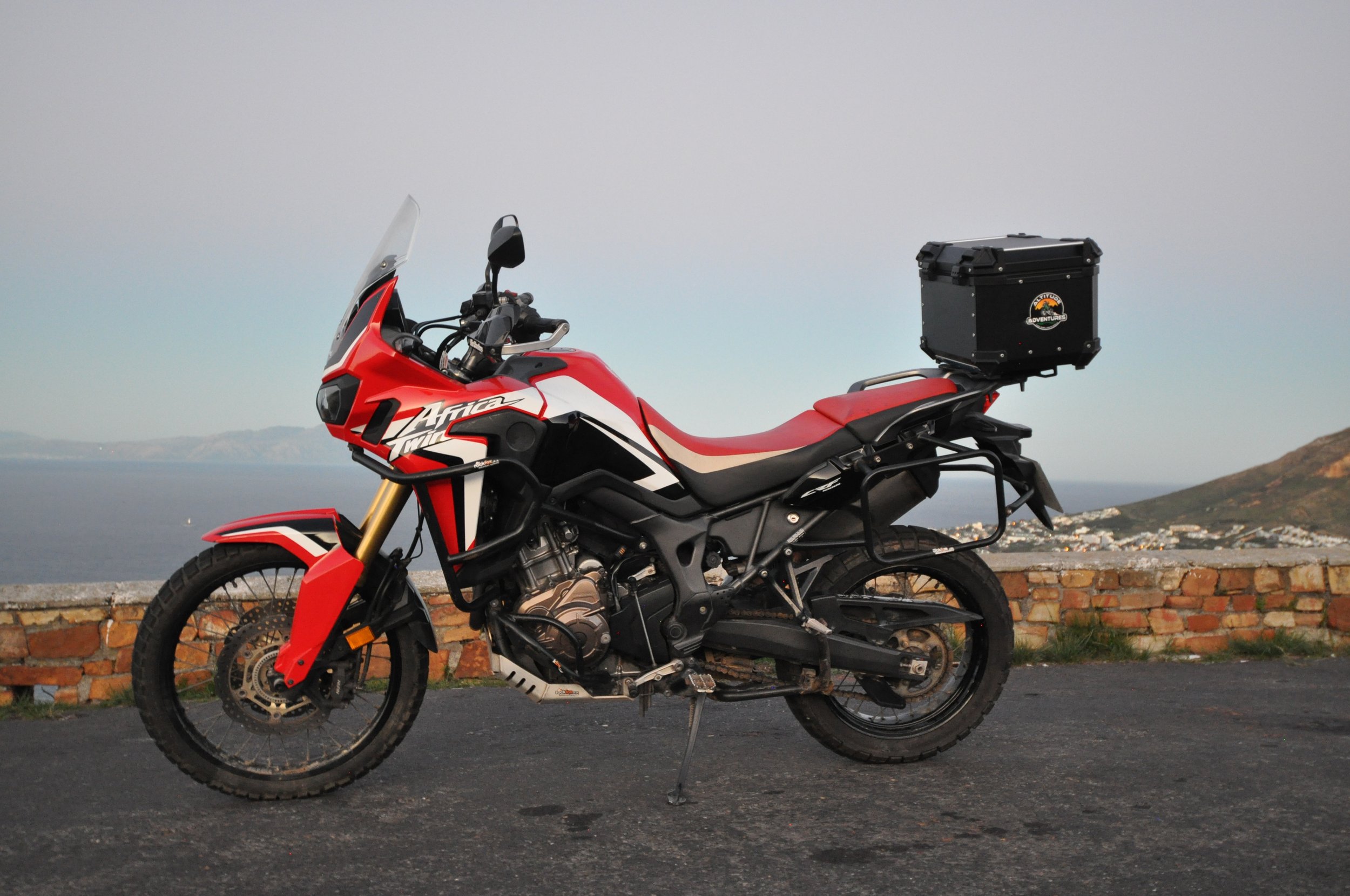 Red and black motorcycle with a top box parked on the side of the road near a brick wall, with a scenic landscape of the water and hills in the background.