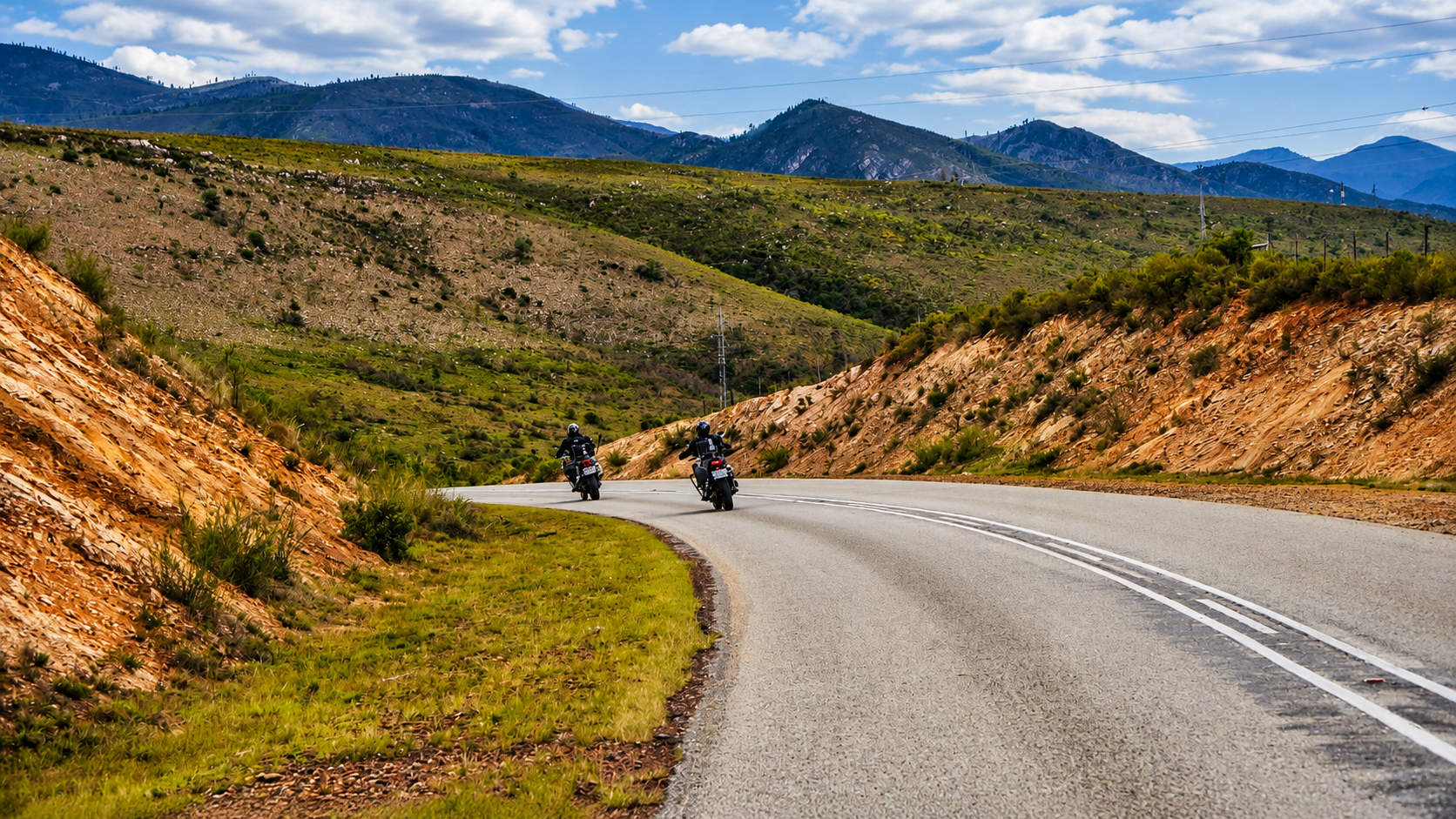 Two motorcyclists riding along a winding road through a mountainous landscape with green hills and rocky slopes.