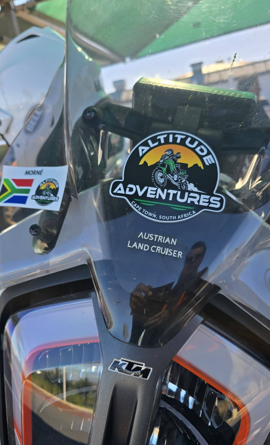 Close-up of a motorcycle windshield with sticker logos, including one for 'Altitude Adventures' in Cape Town, South Africa, describing it as an Austrian land cruiser. Reflection of a person taking the photo can be seen.