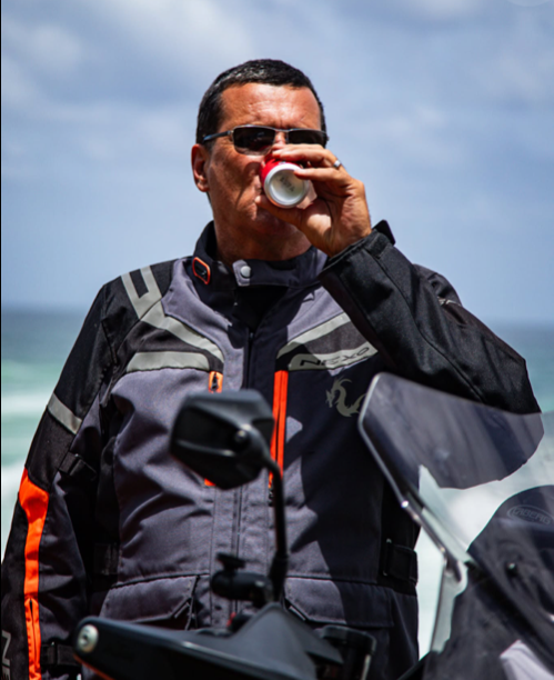Man in motorcycle gear drinking from a can outdoors on a cloudy day.