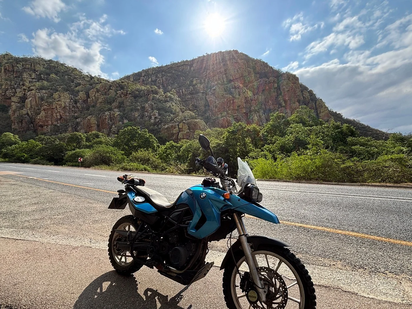 A blue BMW motorcycle parked on the side of a highway with rocky mountain and green vegetation in the background, under a partly cloudy sky.