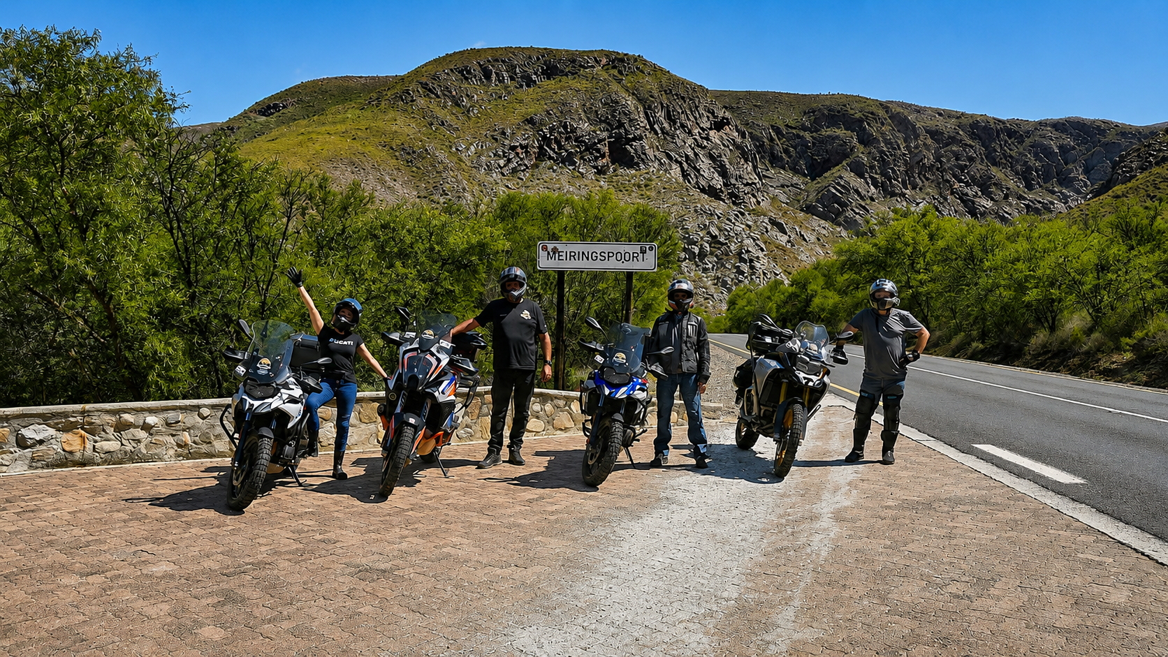 Group of five motorcyclists standing with their bikes on a roadside near a sign that says 'Meiringspoort', with a mountainous landscape and green trees in the background on a sunny day.