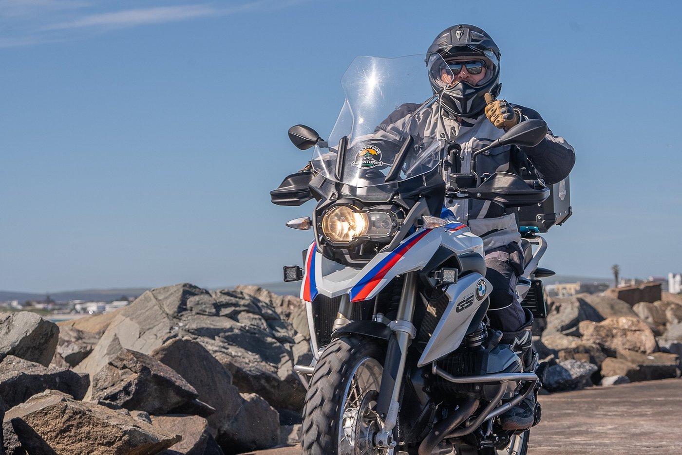 Motorcyclist riding a BMW adventure motorcycle on a rocky shoreline with a clear blue sky.