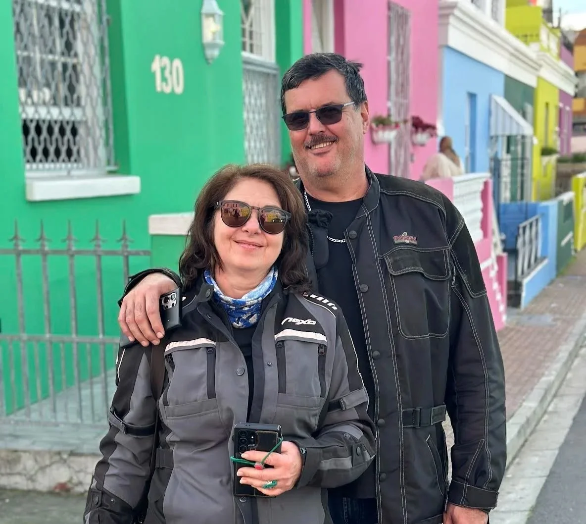 A smiling woman and man in motorcycle gear standing in front of colorful houses on a street. The woman is holding a phone and wearing sunglasses, the man is wearing sunglasses and a black jacket, with his arm around her.