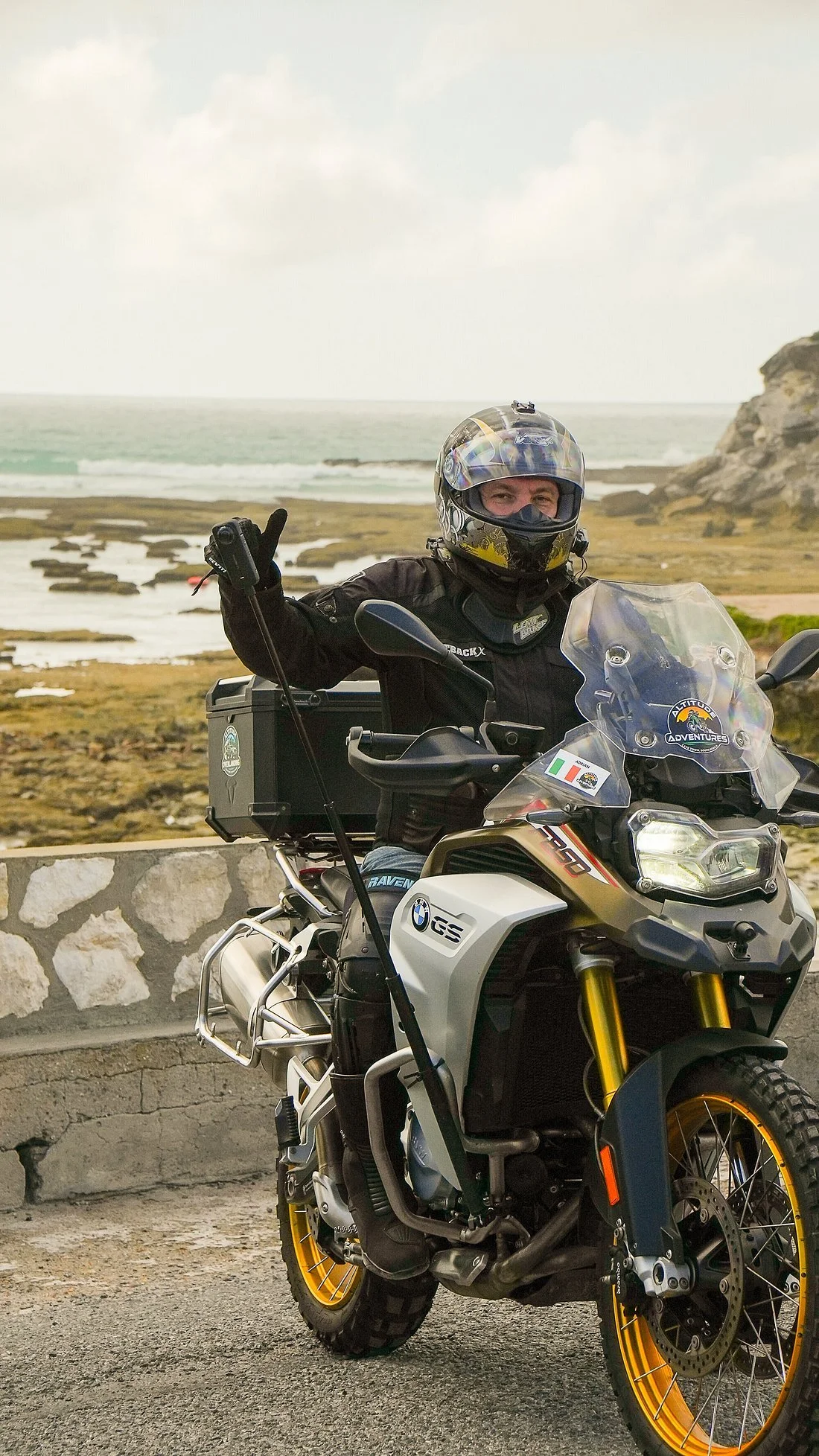 A person wearing a helmet and riding gear sitting on a BMW GS adventure motorcycle near the coast, with water and rocky landscape in the background.
