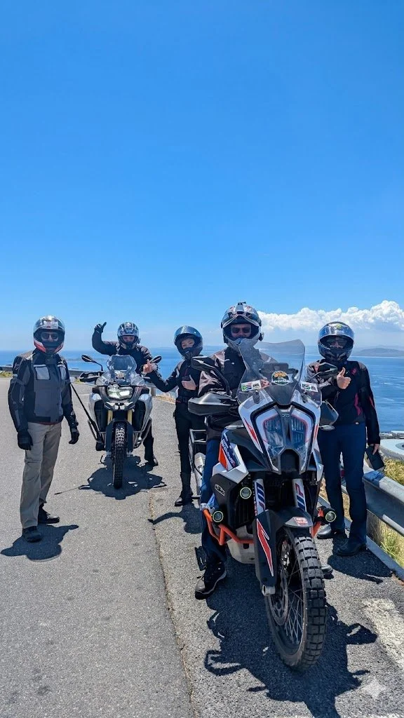 Five people wearing motorcycle gear and helmets, standing with two off-road motorcycles on a coastal road under a clear blue sky.