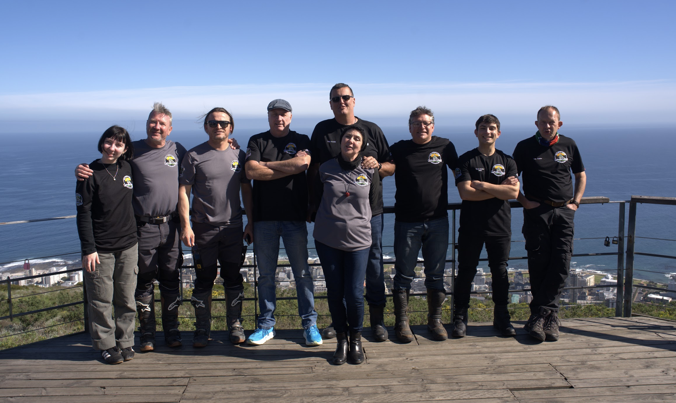 Group of nine people standing on a wooden observation deck against a backdrop of ocean, cityscape, and clear blue sky, with some wearing black or gray shirts with a logo.