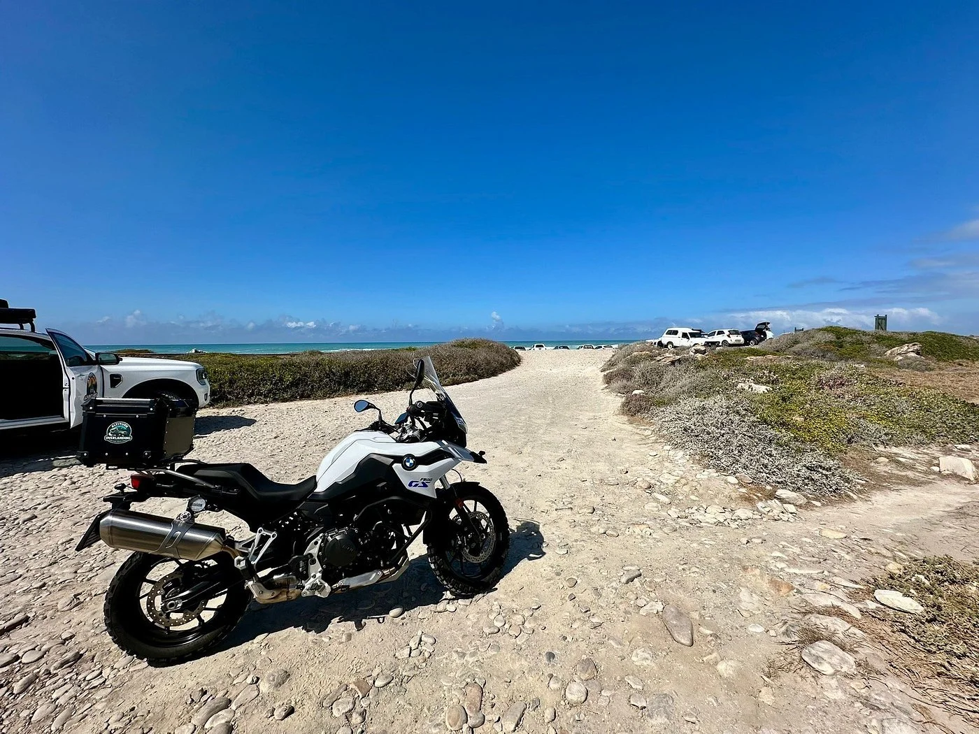 A motorcycle parked on a rocky beach with several vehicles, including an overland vehicle, and the ocean with a blue sky in the background.