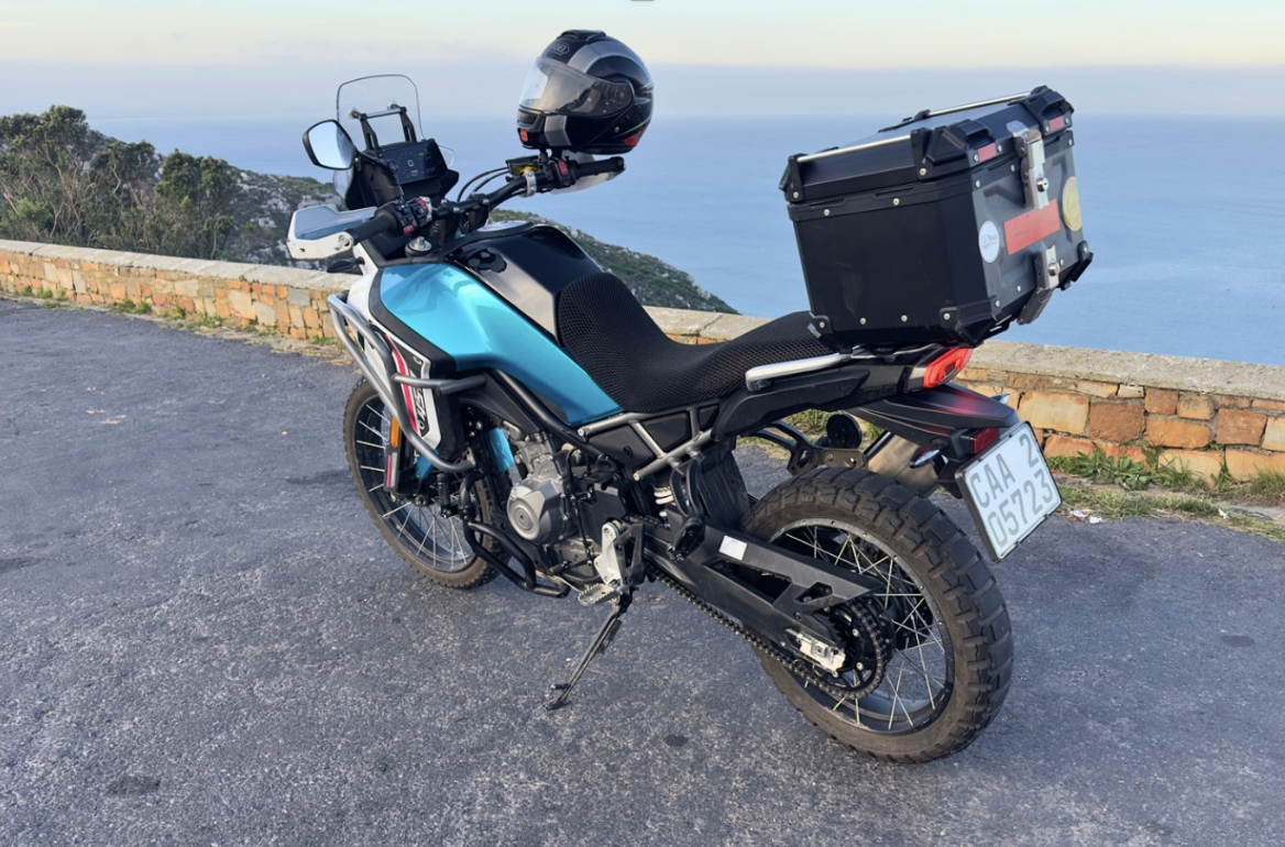 Adventure motorcycle with a top box and helmet, parked on a mountain lookout with ocean view in the background.