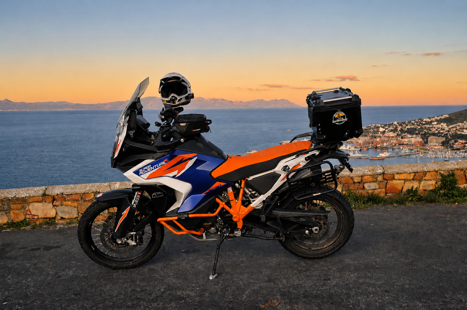 A blue, white, and orange adventure motorcycle parked on the side of a scenic road during sunset, with a coastal city and mountains in the background.