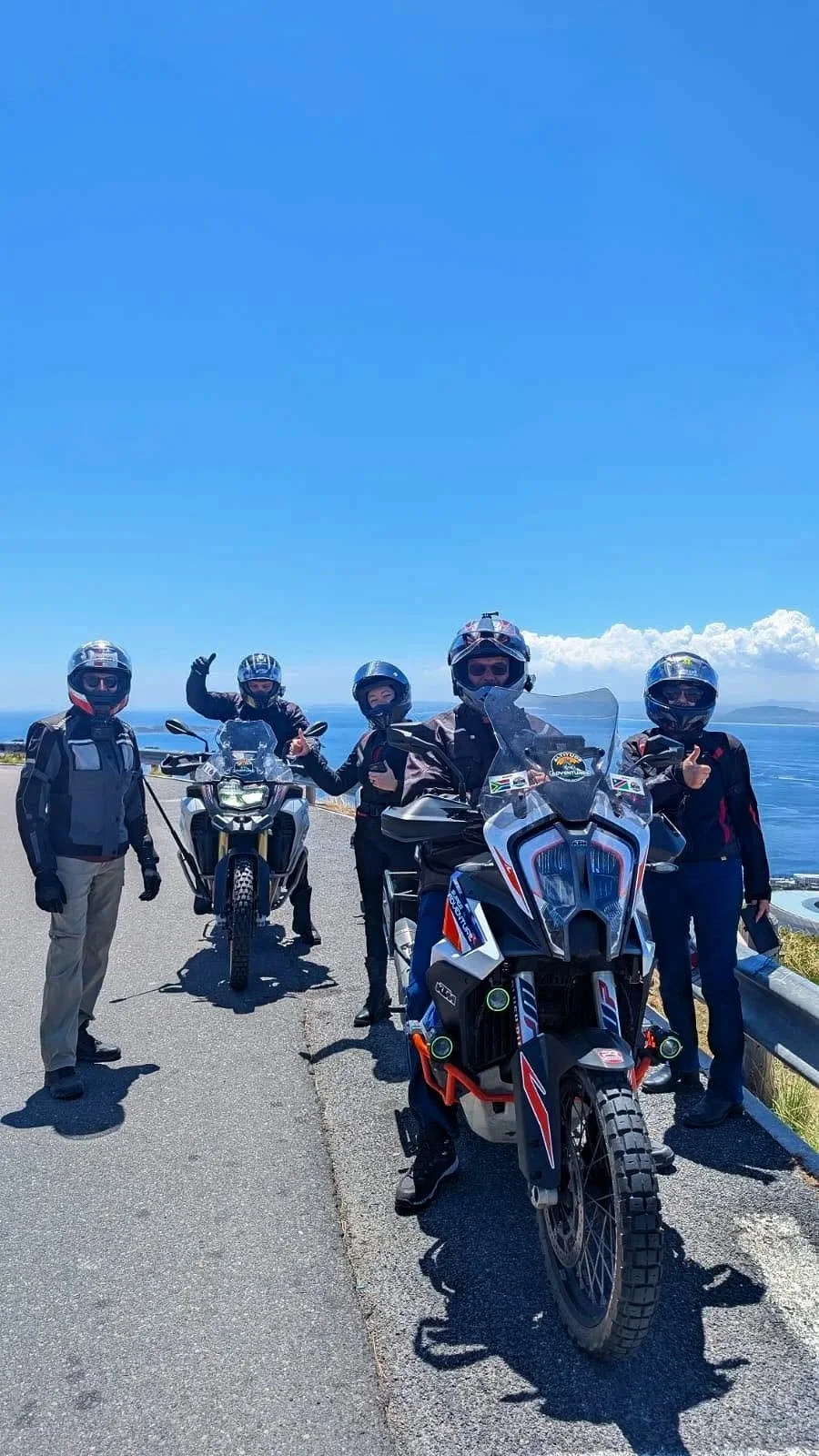 Group of five motorcyclists wearing helmets and protective gear standing outdoors on a road with motorcycles, with ocean and sky in the background.
