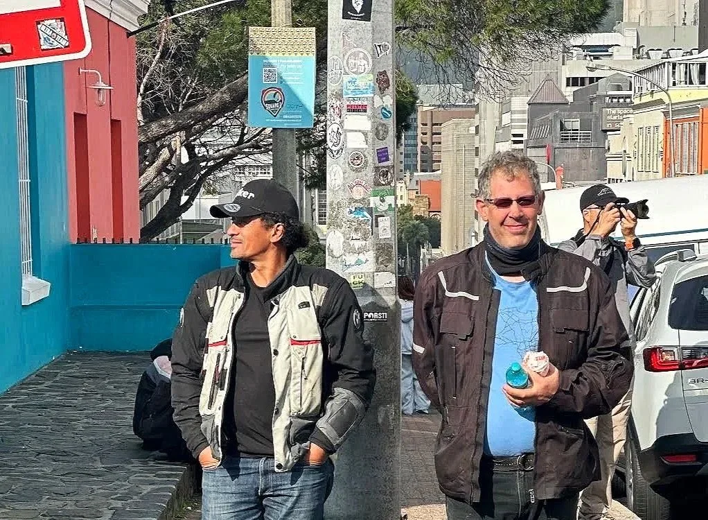 Two men standing outside on a city street, one holding a water bottle, the other with hands in pockets, with a photographer taking pictures in the background in Bo-kaap Cape Town South Africa. Motorcycle tour guides altitude adventures