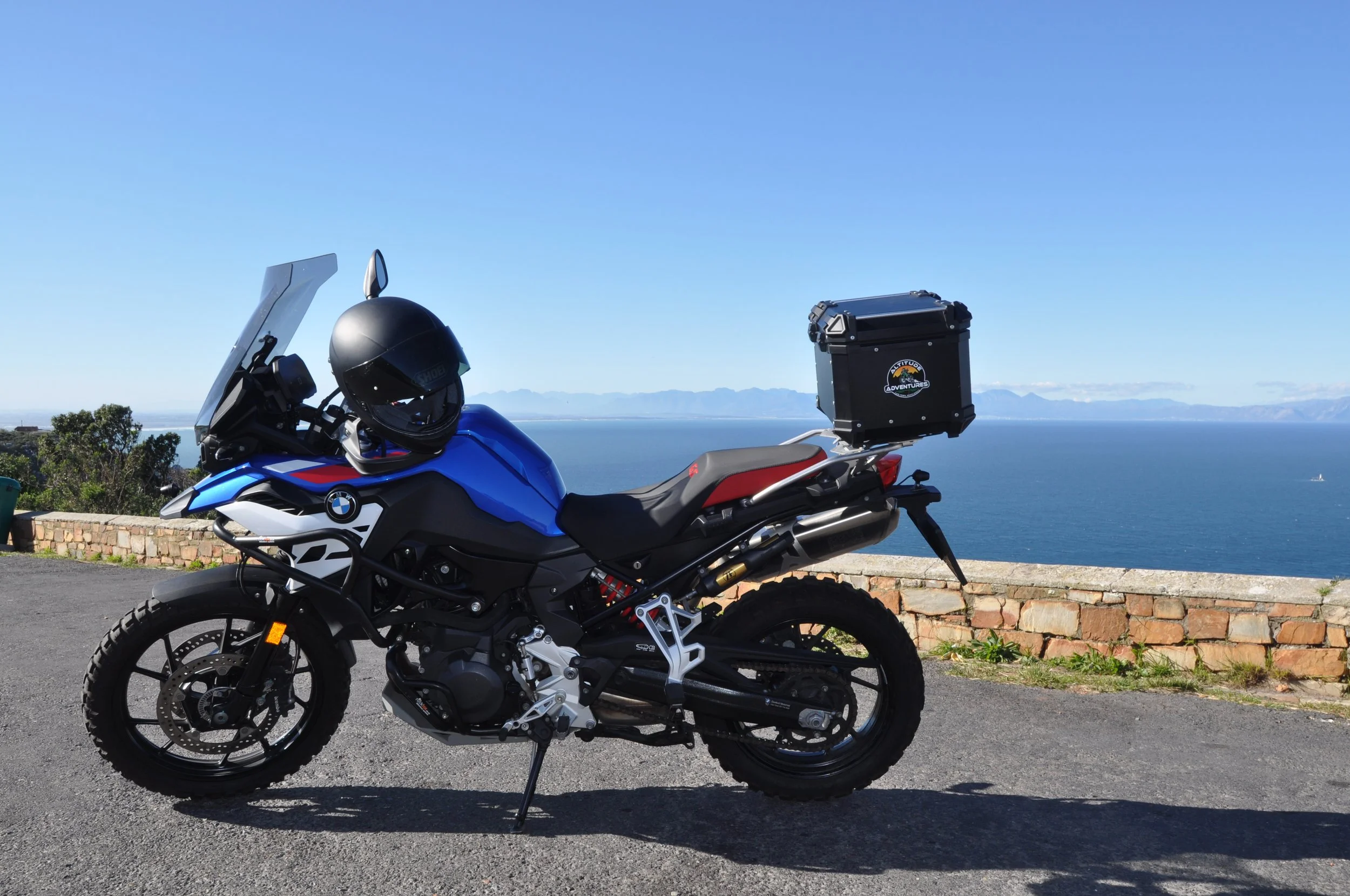 Blue and black BMW adventure motorcycle with a black helmet on the handlebar, parked on a road near a brick wall overlooking an ocean, with mountains in the distance and a clear blue sky.