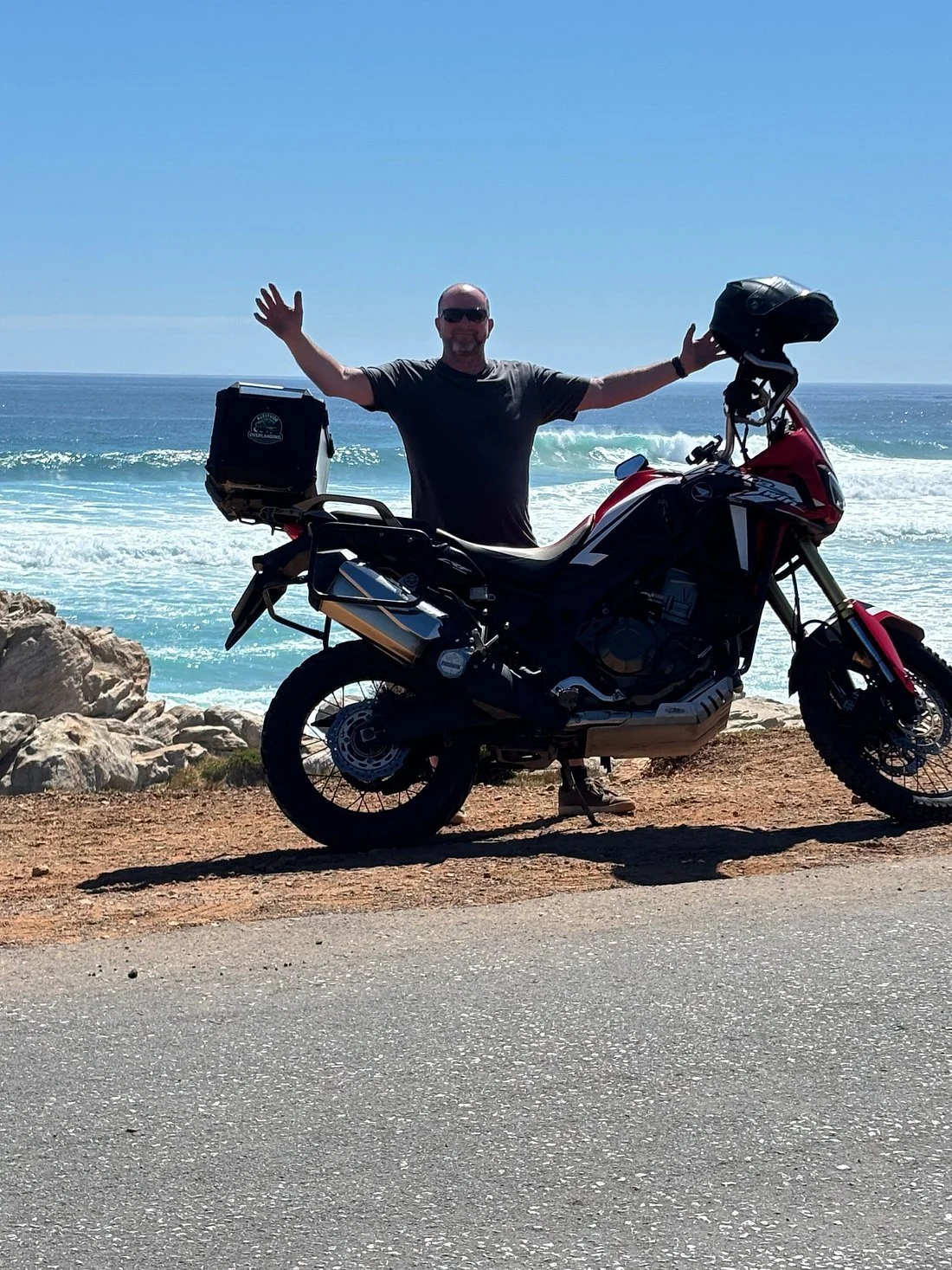 A man standing next to a red and black motorcycle on a rocky beach with waves and ocean in the background, raising his arms. Misty Cliffs, Cape Town. 
