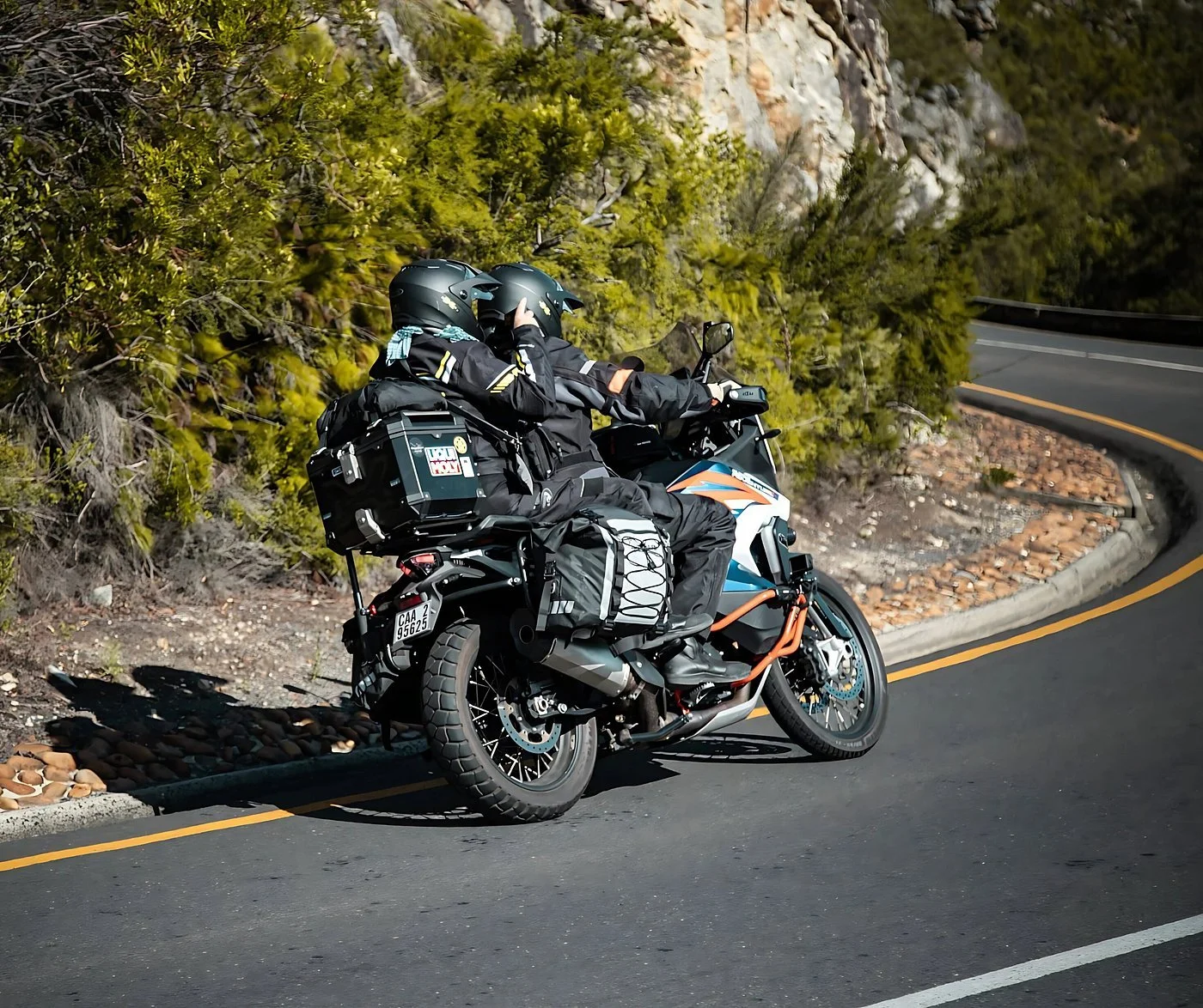 Two motorcyclists wearing helmets riding on a winding mountain road with luggage strapped to their motorcycle, surrounded by green trees and rocky terrain.