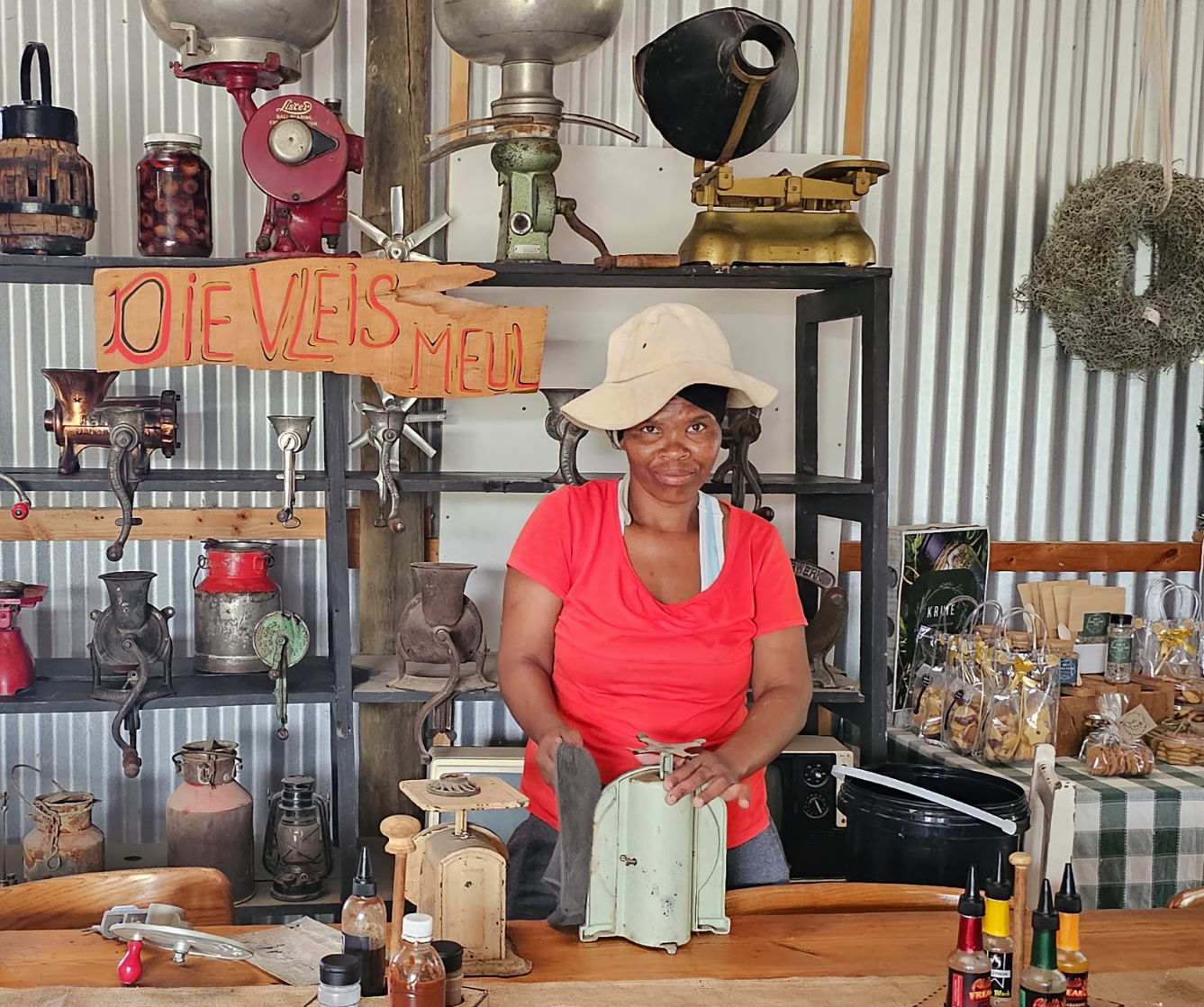 Woman in a red shirt and wide-brimmed hat standing behind a counter with various vintage kitchen items and kitchenware on shelves and tables around her.