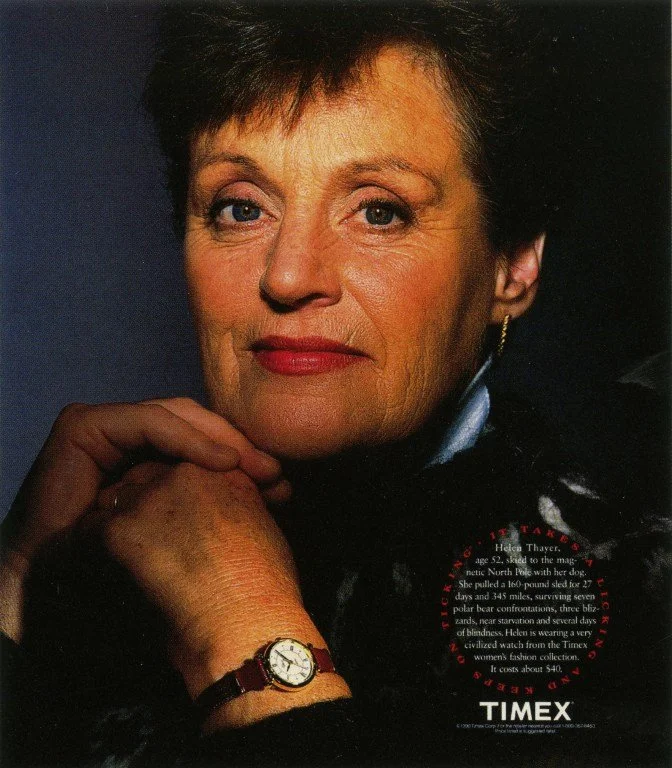 Close-up portrait of Helen Thayer, a mature woman with short dark hair, wearing red lipstick, jewelry, and a watch, with a watch magazine in the background.