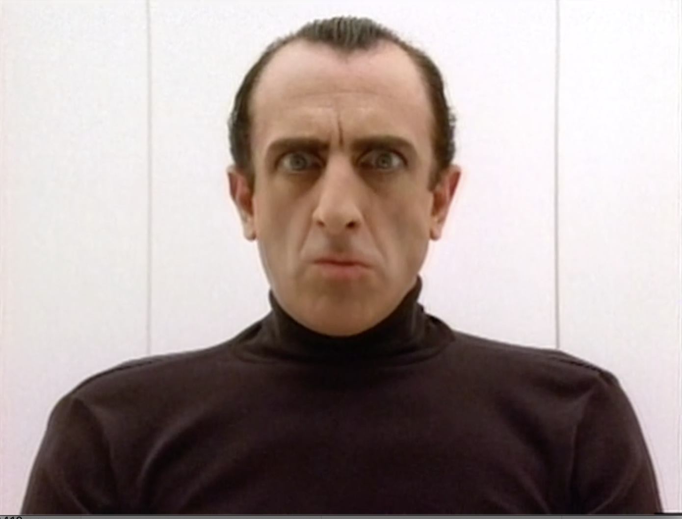 A man with short dark hair, wearing a black turtleneck, sitting against a plain white wall, with a serious expression.