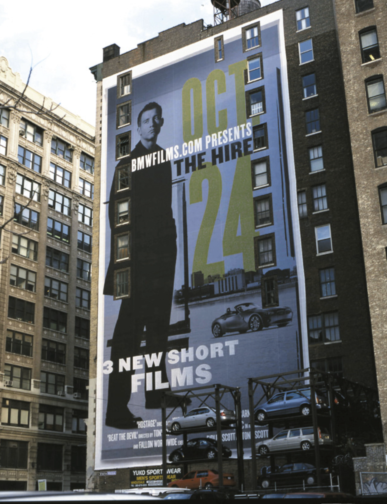 Large billboard on a building advertising a film release. The poster features a man in a dark coat with a cityscape and a car in the background. Text reads "OCT 24," "bmwfilms.com presents," "The Hire," and "3 new short films."