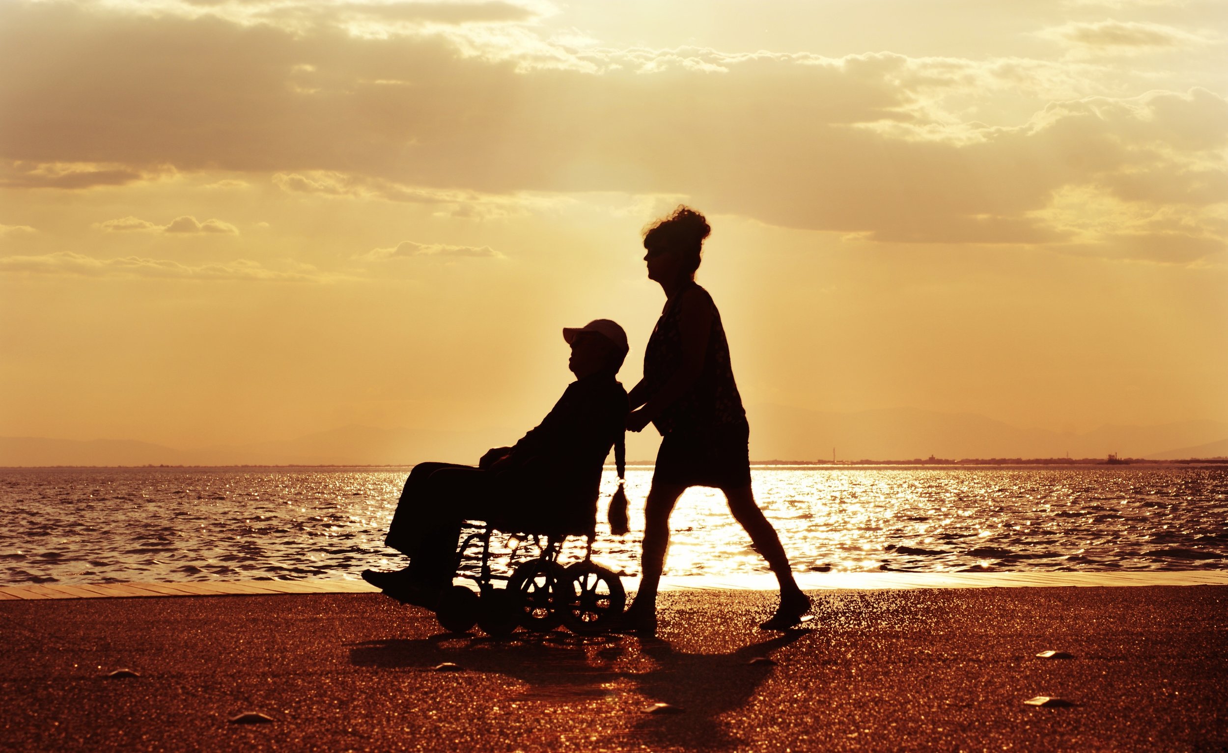 A silhouette of a person riding a wheelchair by the water, with another person standing behind them, during a golden sunset with cloudy sky.