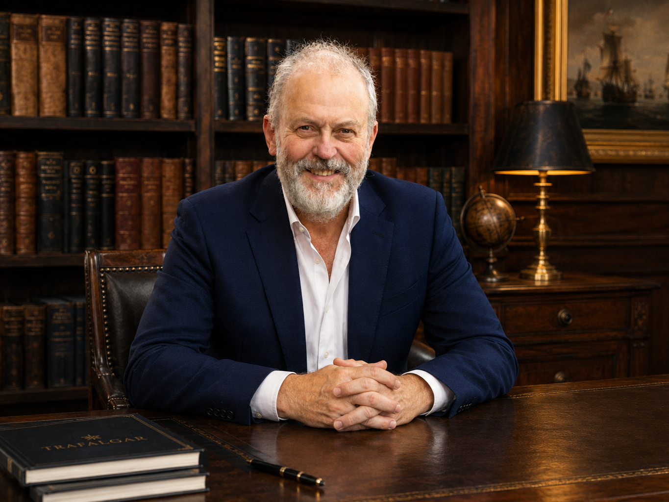 An older man with white hair and beard, wearing a navy blazer and white shirt, sitting at a wooden desk in a library with dark wood bookshelves filled with old books, a black table lamp, a framed painting, and a globe in the background.