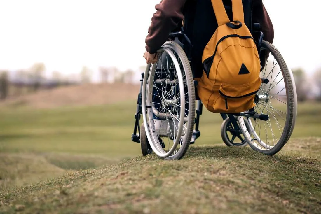 Person in a wheelchair with a yellow backpack sitting outdoors on grassy terrain.