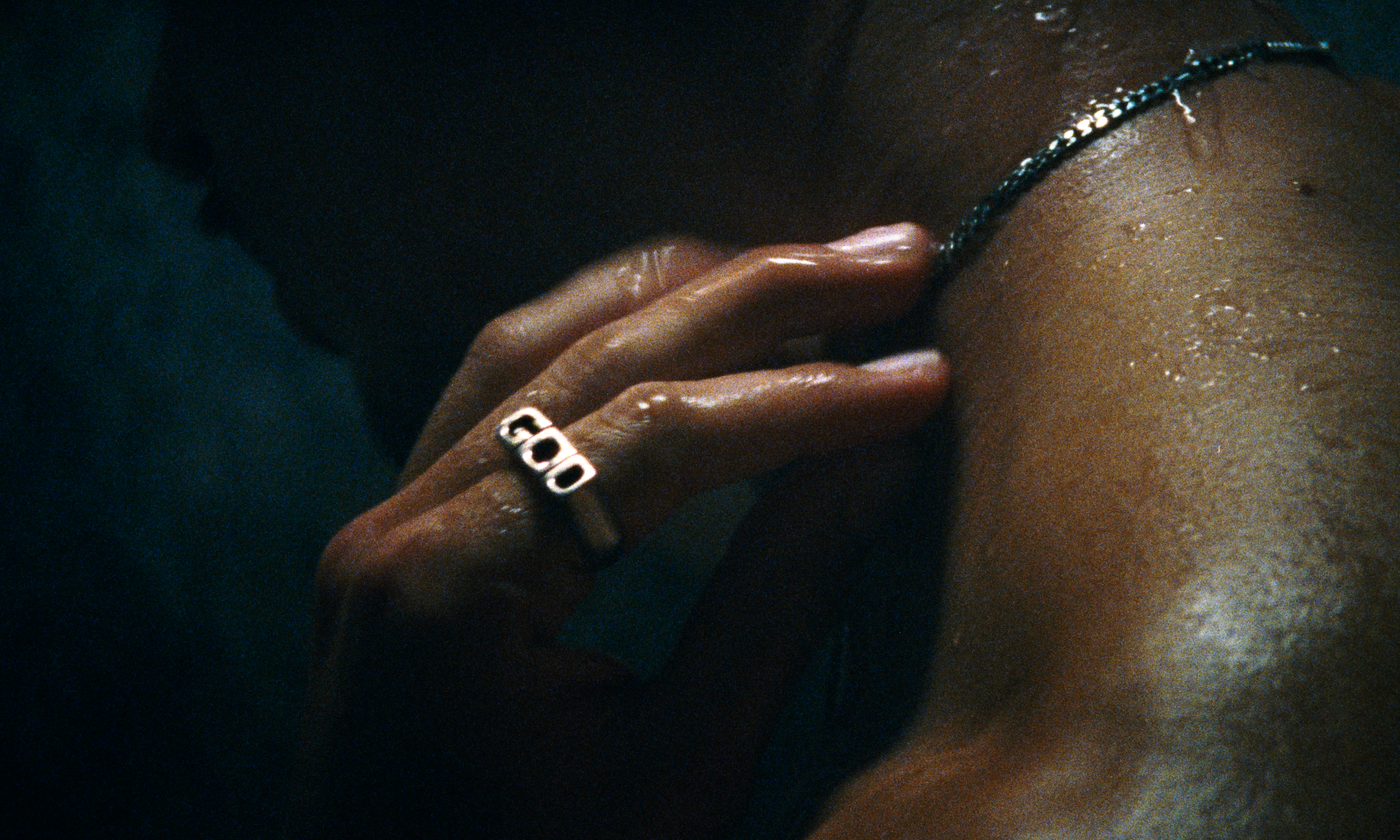 Close-up of a person's hand wearing a ring with the word 'GOD,' touching their neck, with a necklace visible on their skin.