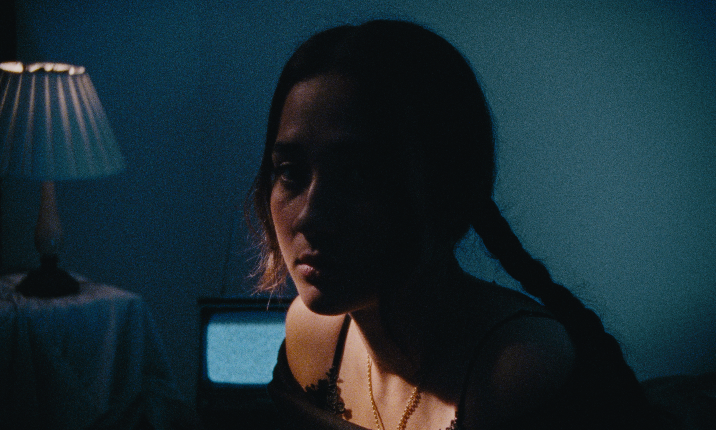 Dark-haired woman with braided hair wearing a black top and gold necklace, sitting in a dimly lit room with a blue wall, a lamp with a patterned shade on her left, and a television screen behind her.