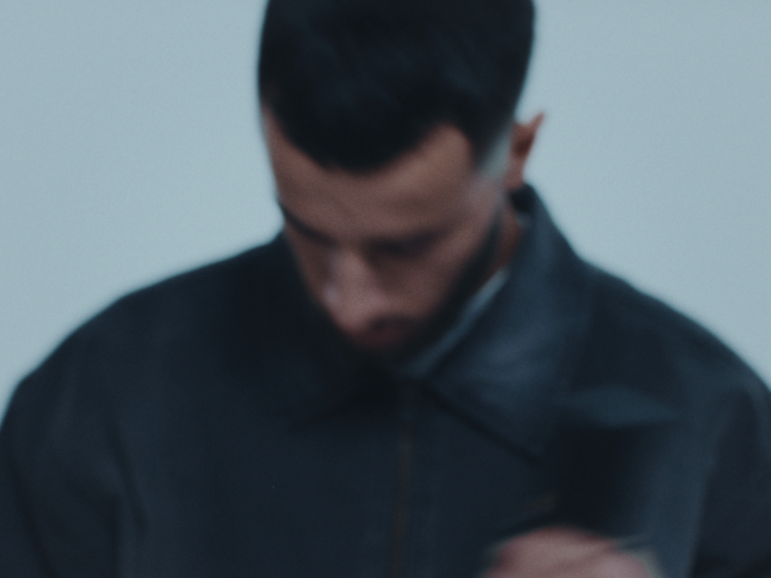 Blurry close-up photo of a person with short dark hair, wearing a dark jacket, with a plain light-colored background.