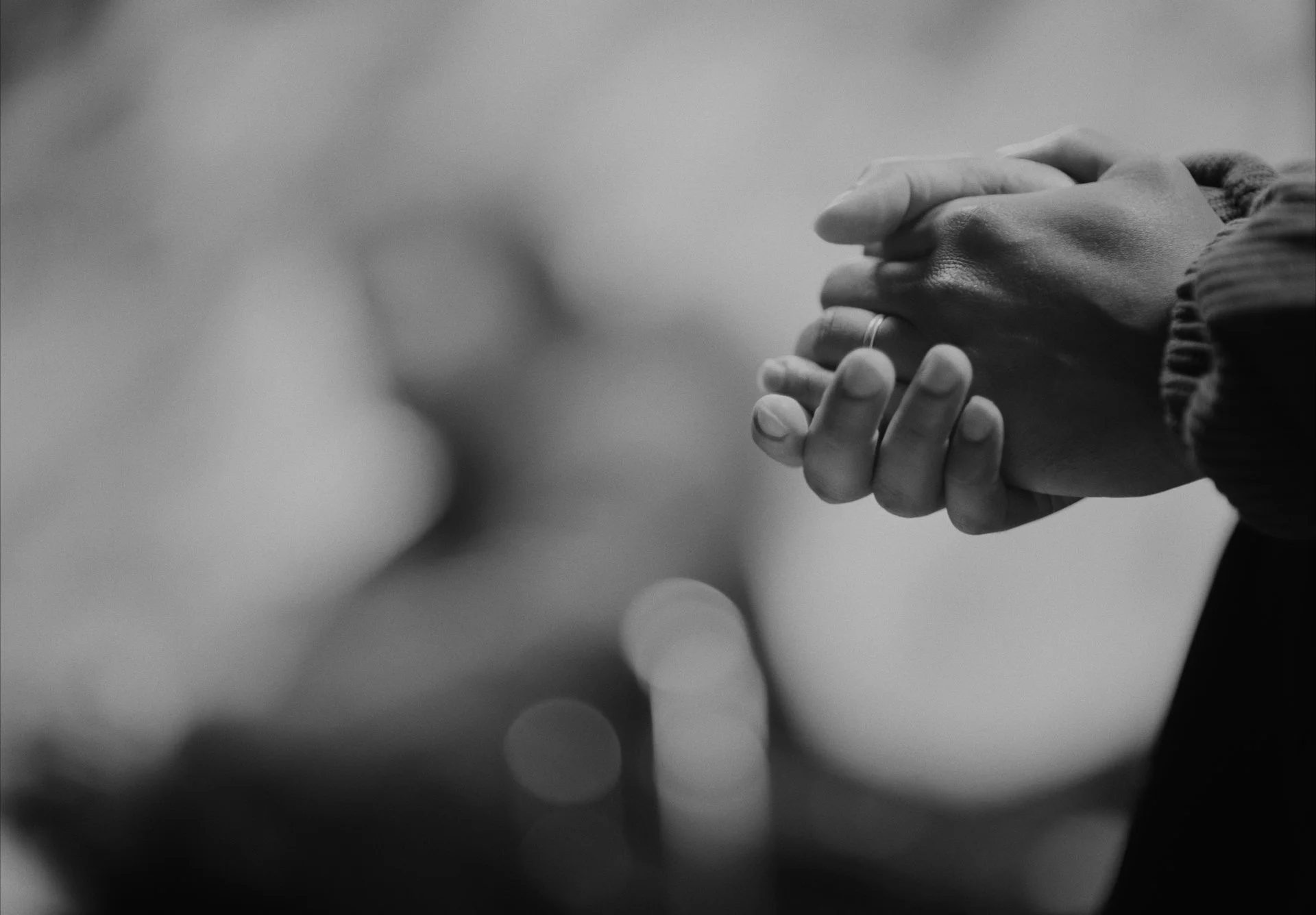 Close-up of two hands clasped together, one with a wedding ring, in a praying or contemplative pose, in black and white.
