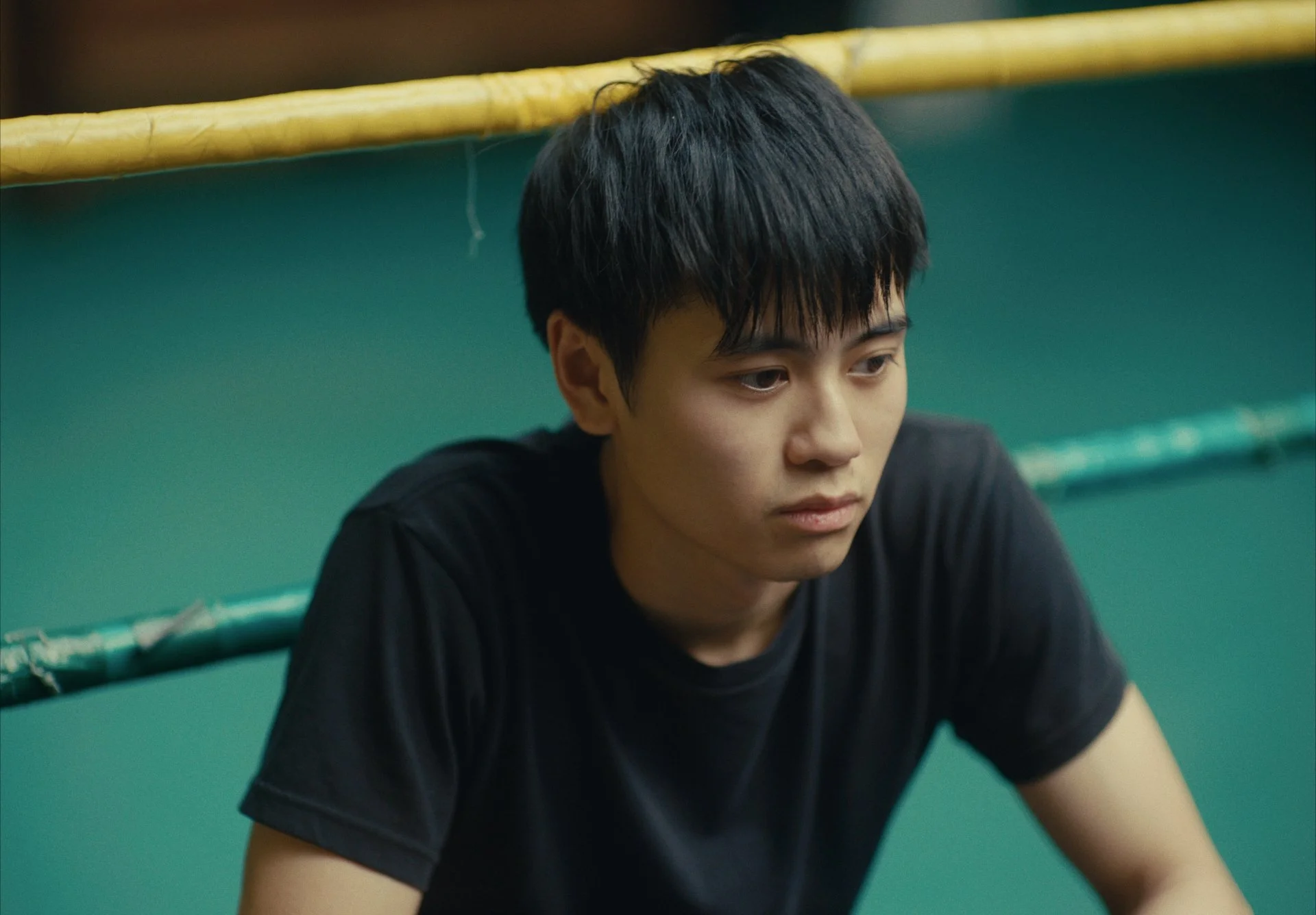 Young man with black hair in a black t-shirt sitting near a green wall with yellow piping, looking down with a serious expression.
