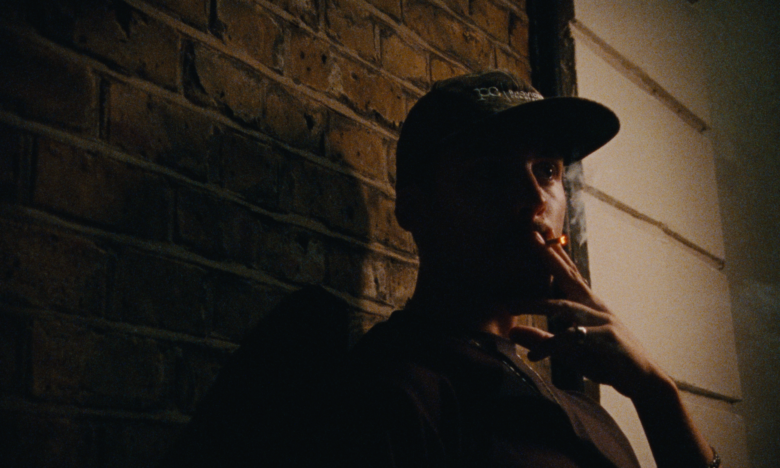 A person wearing a baseball cap and smoking a cigarette, leaning against a brick wall in a dimly lit room.
