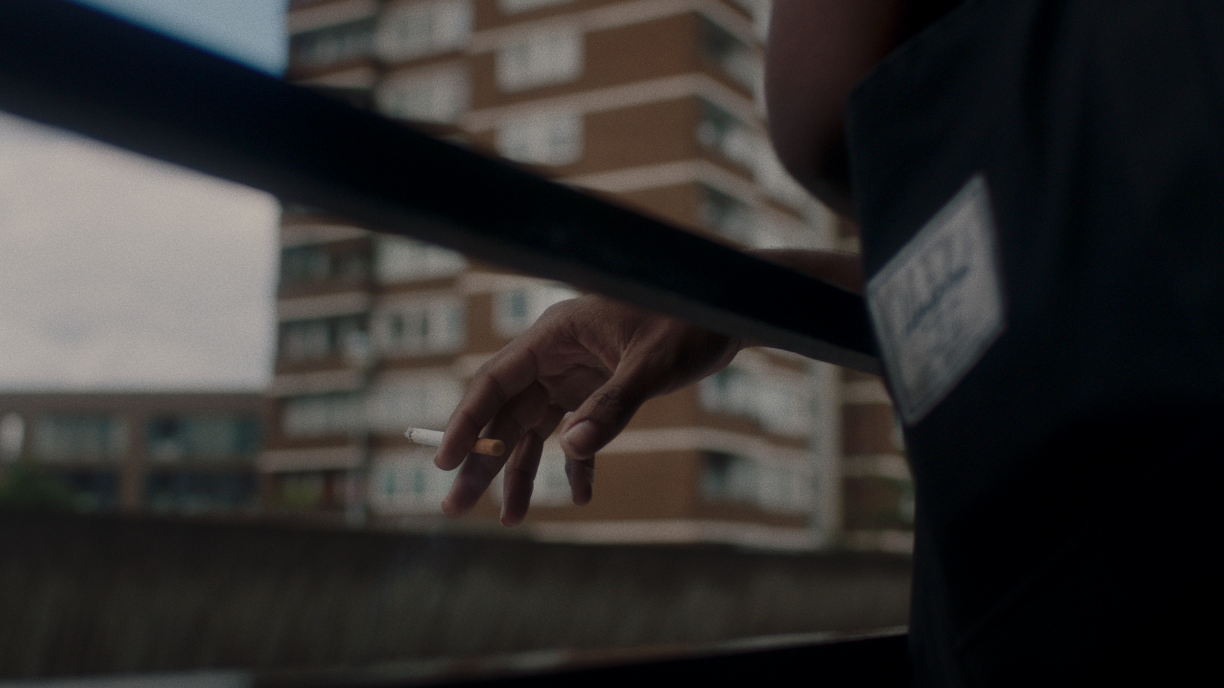 Close-up of a person's hand holding a cigarette, with a blurred background of apartment buildings.