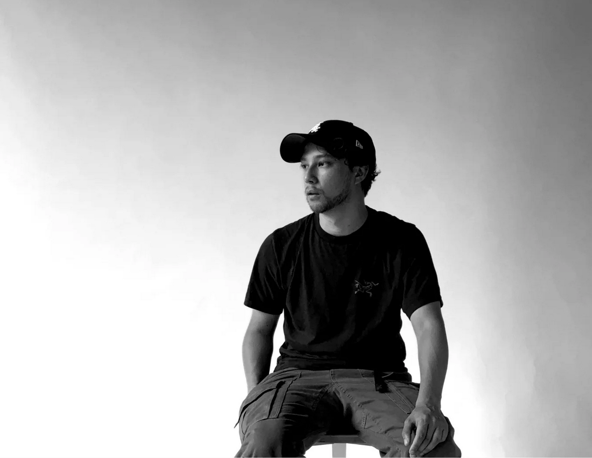A man wearing a black baseball cap and t-shirt sitting on a stool against a gradient background.