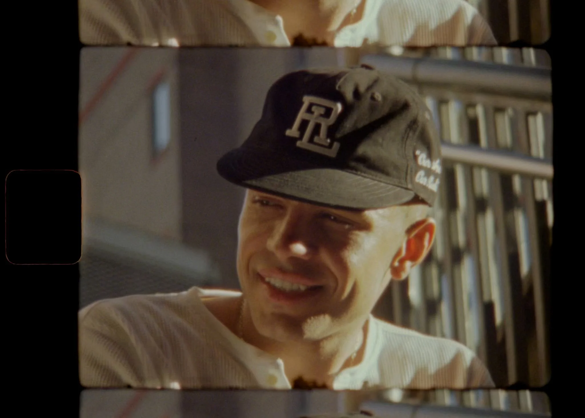 A man wearing a gray baseball cap with a white letter 'R' on the front, smiling, in front of bookshelves.