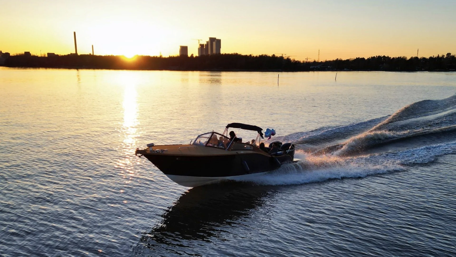 A motorboat speeding across a body of water during sunset, with a city skyline of high-rise buildings and smokestacks in the background.
