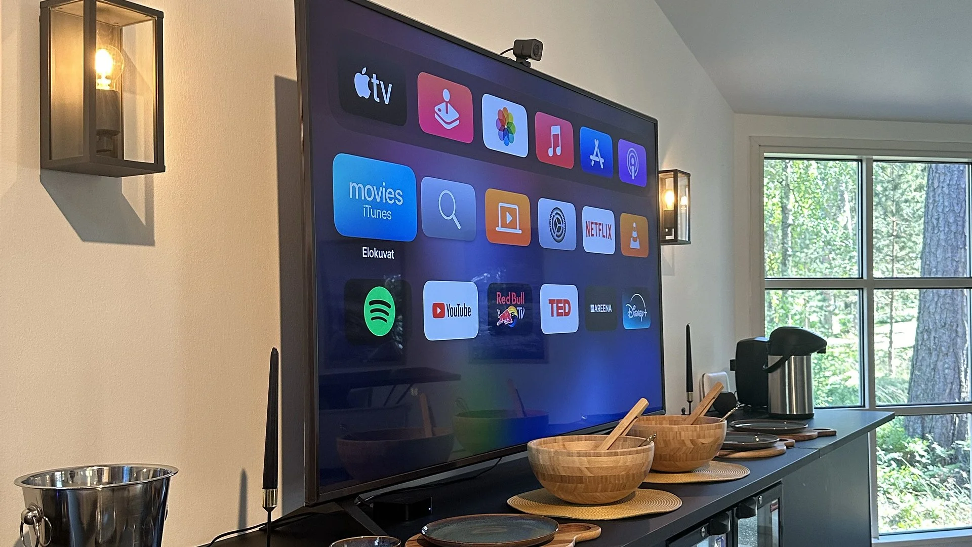 Living room with a large flat-screen TV displaying streaming apps, above a sideboard with wooden bowls, a coffee pot, and social media icons. Wall-mounted lights and a window showing trees are visible.