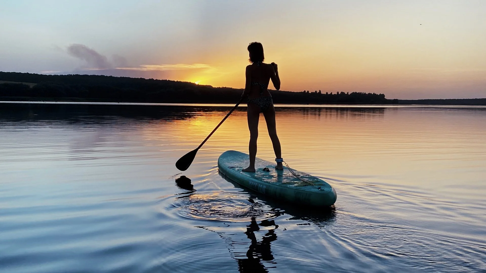 Person paddleboarding on a calm lake during sunset with a silhouette of trees in the background.