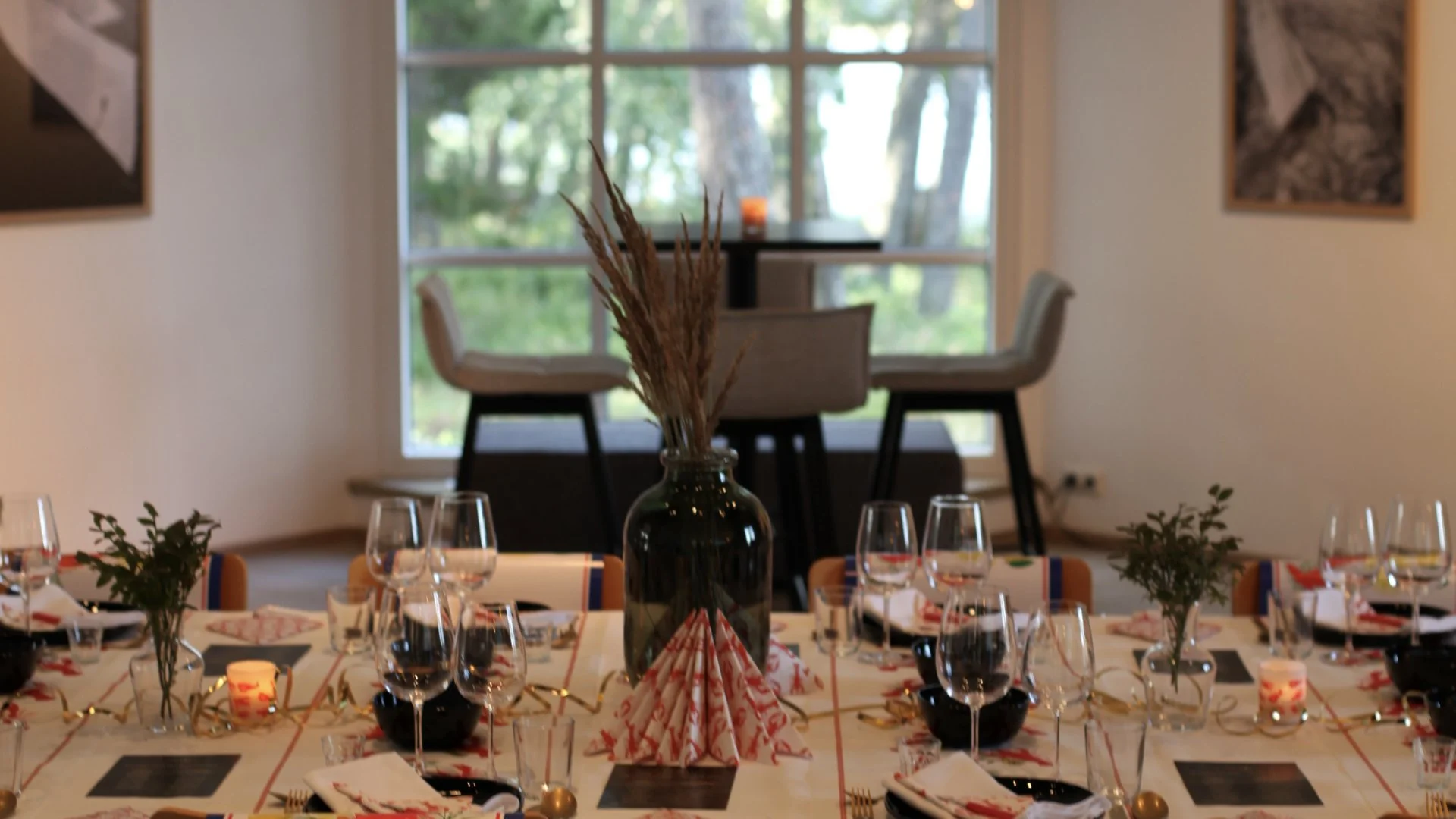 A dining table set for a celebration with a centerpiece consisting of a dark bottle holding dried wheat stalks. The table is decorated with small candles, green plants, and festive napkins. In the background, a window with a view of trees and an inte