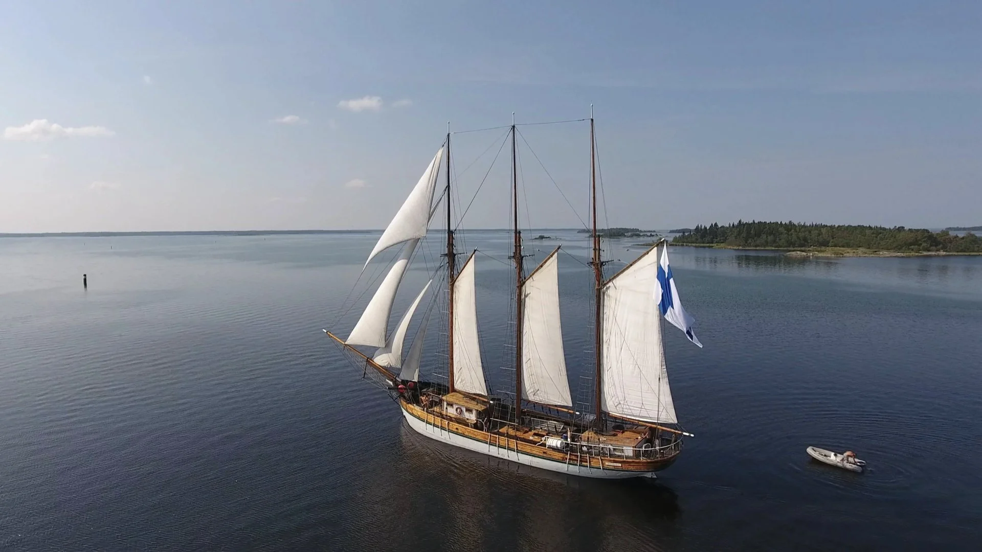 A large traditional sailing ship with white sails and a Finnish flag, floating on calm water near a forested shoreline under a clear sky.