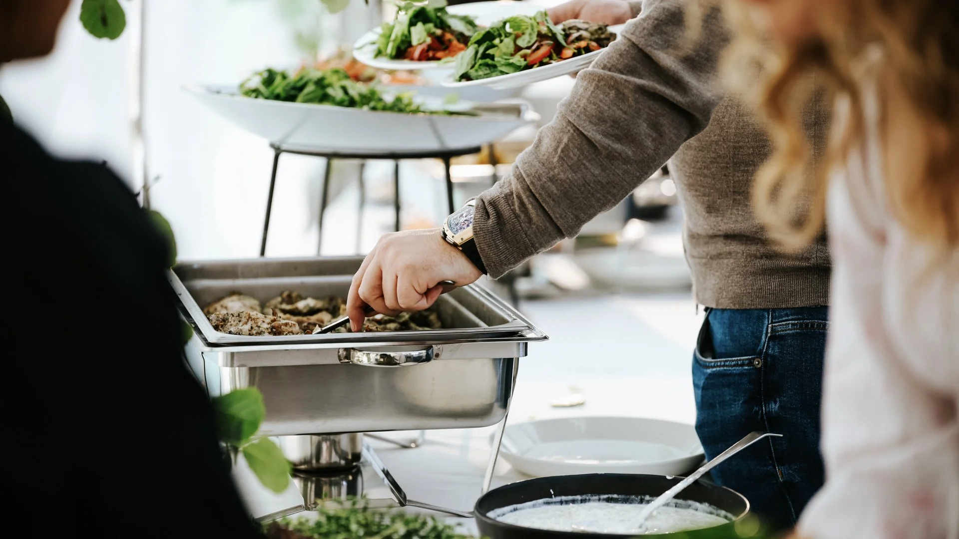 Person serving food from a chafing dish at a buffet with salads and vegetables on display.