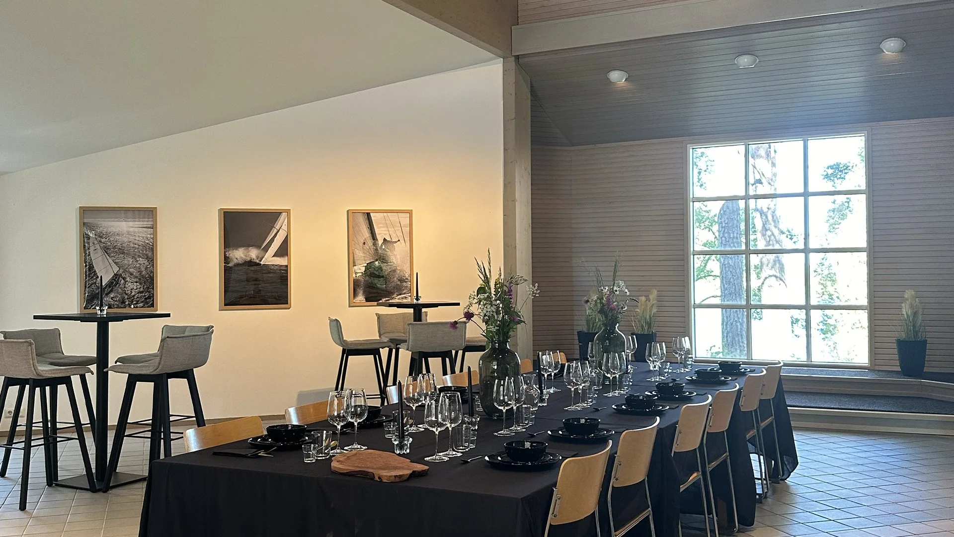 Interior of a dining area with a long table set with glassware, black bowls, and vases with flowers, next to large windows showing trees outside. Small round tables with chairs are in the background, and framed photographs of sailboats hang on the wa