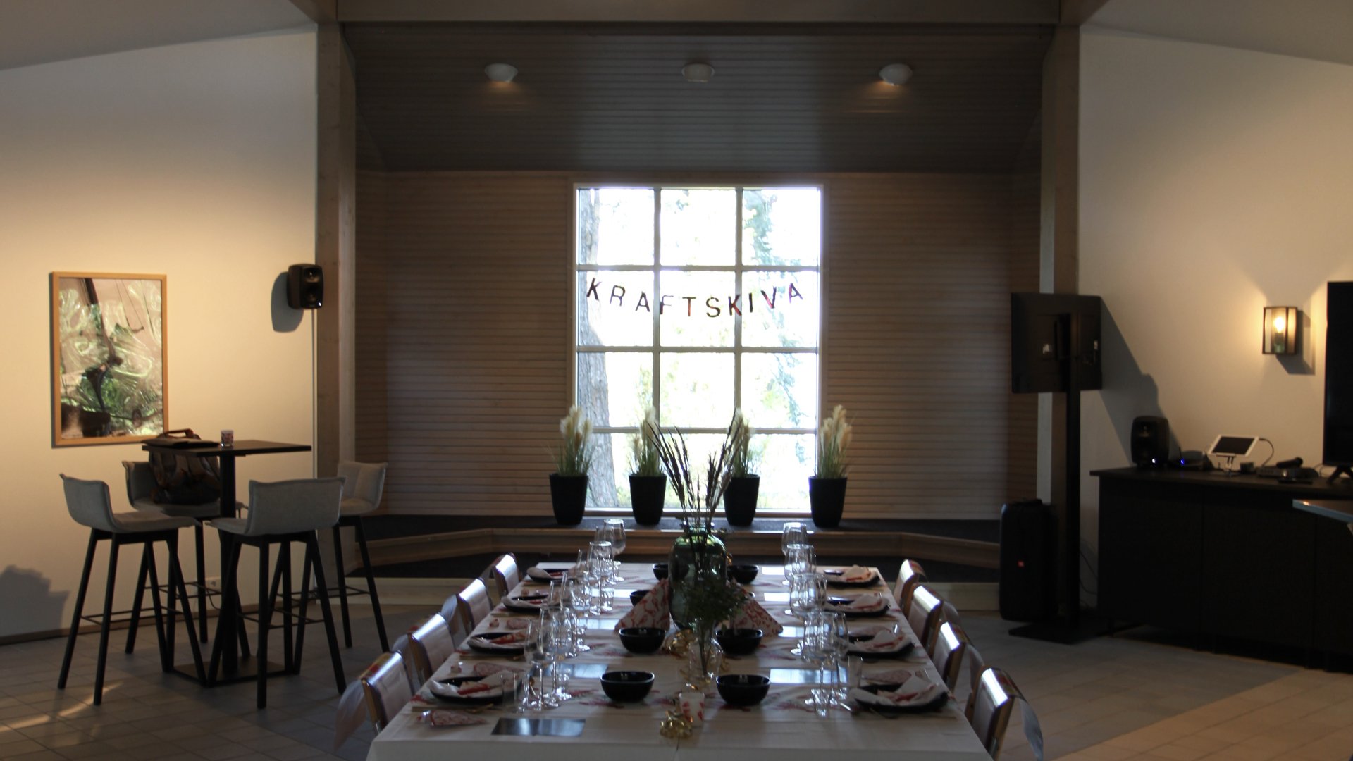Decorated dining area with a long table set for a meal, featuring white tablecloths, glassware, and bowls. Behind the table, there are four black potted plants and a large window with sunlight filtering through. To the left, a high table with four ch