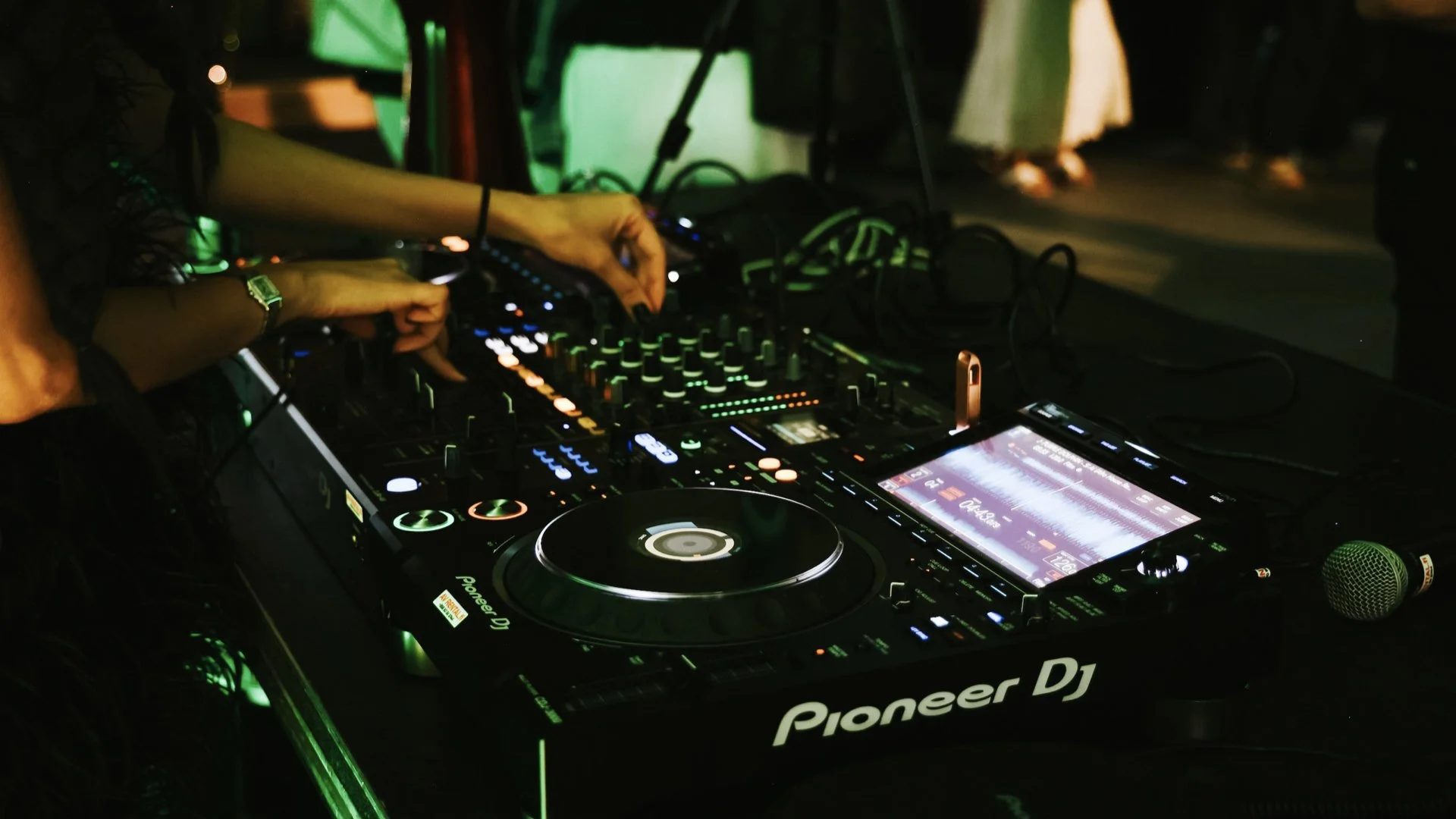 A DJ setup with Pioneer DJ equipment, including a mixer, turntables, and a touchscreen controller, illuminated with green and orange lighting, with a microphone nearby.